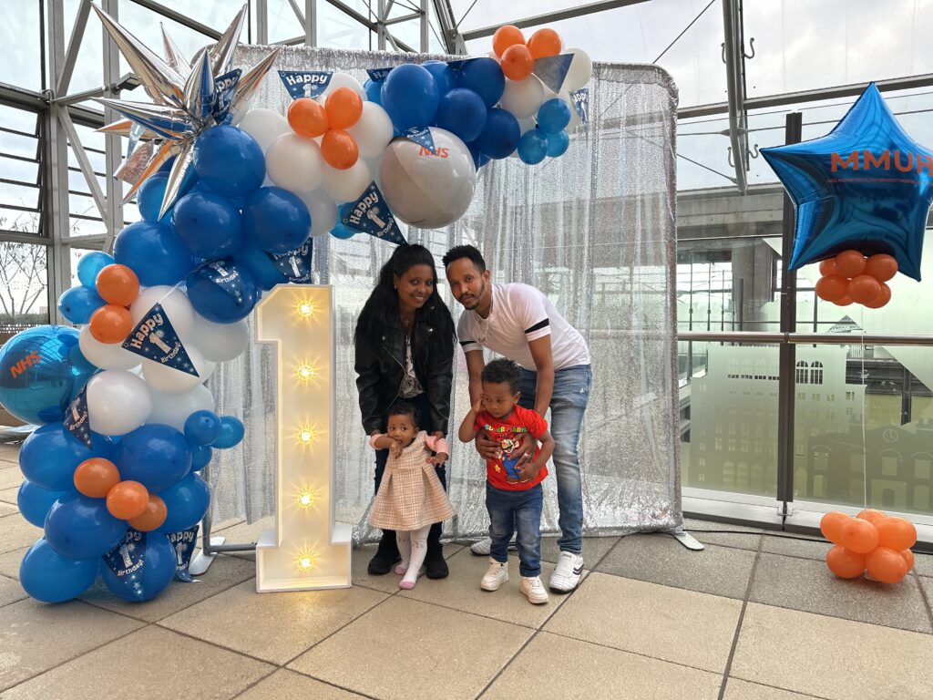 An image of a family with their two children, in front of a illuminated number 1 and birthday balloons.